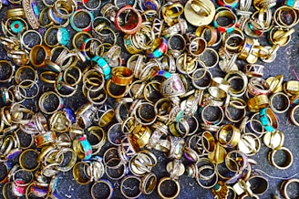 A large collection of various rings scattered on a dark surface. The rings display a variety of designs, with some featuring turquoise stones, intricate patterns, and metallic finishes. Their colors range from gold and silver to bright blues and reds, creating an eclectic and vibrant assortment.
