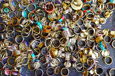 A large collection of various rings scattered on a dark surface. The rings display a variety of designs, with some featuring turquoise stones, intricate patterns, and metallic finishes. Their colors range from gold and silver to bright blues and reds, creating an eclectic and vibrant assortment.