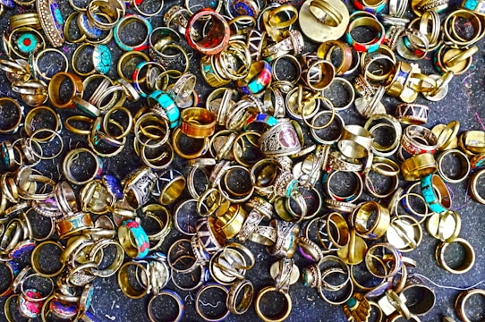 A large collection of various rings scattered on a dark surface. The rings display a variety of designs, with some featuring turquoise stones, intricate patterns, and metallic finishes. Their colors range from gold and silver to bright blues and reds, creating an eclectic and vibrant assortment.