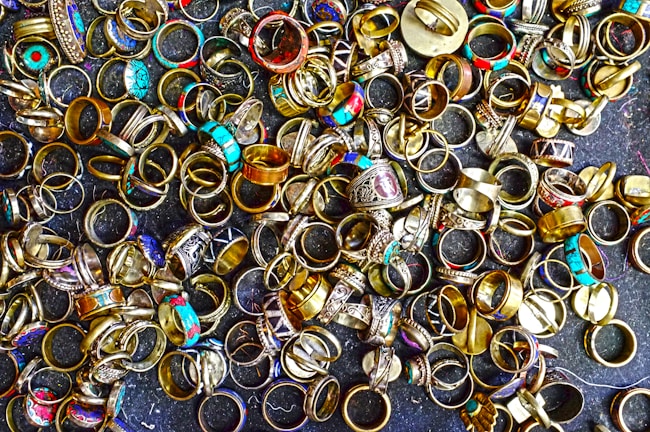 A large collection of various rings scattered on a dark surface. The rings display a variety of designs, with some featuring turquoise stones, intricate patterns, and metallic finishes. Their colors range from gold and silver to bright blues and reds, creating an eclectic and vibrant assortment.