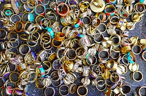 A large collection of various rings scattered on a dark surface. The rings display a variety of designs, with some featuring turquoise stones, intricate patterns, and metallic finishes. Their colors range from gold and silver to bright blues and reds, creating an eclectic and vibrant assortment.