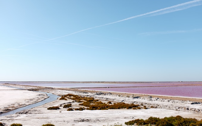 A vast, open landscape with a series of parallel salt pans filled with pink water, surrounded by white, sandy banks and sparse vegetation. A small stream meanders through the foreground, set against a clear blue sky with faint contrails.