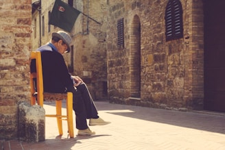 An older man is sitting pensively on a yellow wooden chair in a sunlit narrow street. His legs are crossed, and he wears a hat and glasses. The background features rustic stone buildings with dark shutters and a flag hanging above.