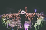 A performer on stage is facing a large, energetic crowd. The audience is engaged, with many people raising their hands and cheering. The setting appears to be a nighttime concert, with stage lights illuminating the scene. Photographers are capturing the event from the sidelines.