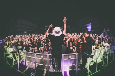 A performer on stage is facing a large, energetic crowd. The audience is engaged, with many people raising their hands and cheering. The setting appears to be a nighttime concert, with stage lights illuminating the scene. Photographers are capturing the event from the sidelines.