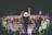 A performer on stage is facing a large, energetic crowd. The audience is engaged, with many people raising their hands and cheering. The setting appears to be a nighttime concert, with stage lights illuminating the scene. Photographers are capturing the event from the sidelines.
