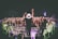 A performer on stage is facing a large, energetic crowd. The audience is engaged, with many people raising their hands and cheering. The setting appears to be a nighttime concert, with stage lights illuminating the scene. Photographers are capturing the event from the sidelines.