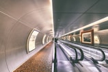 A futuristic tunnel with a moving walkway stretches forward. The tunnel is lined with framed artwork lit from above and large, curved advertisements on the opposite wall. The ceiling and walls have a sleek, metallic design, and a patterned carpet runs along the floor beside the walkway.
