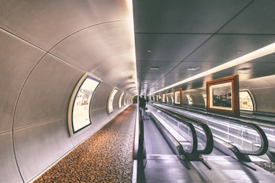A futuristic tunnel with a moving walkway stretches forward. The tunnel is lined with framed artwork lit from above and large, curved advertisements on the opposite wall. The ceiling and walls have a sleek, metallic design, and a patterned carpet runs along the floor beside the walkway.