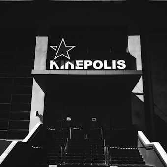 A black and white photograph of a building entrance with a lit sign displaying the word KINEPOLIS in white capital letters next to a stylized star logo. The composition includes stairs leading up to the entrance, which is framed by large, rectangular architectural elements.