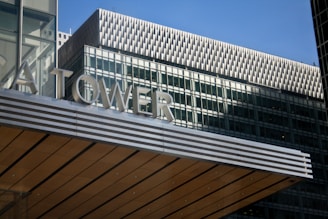 The image features a modern building with a prominent sign reading 'TOWER'. The architecture includes a striped pattern on a horizontal surface, likely part of a canopy or entrance. The background showcases a high-rise office building with a grid of windows and textured upper facade.