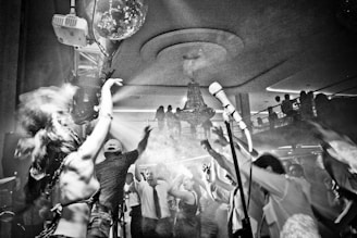 A lively party scene with a crowd of people dancing enthusiastically. The setting is a club or event venue, featuring a large chandelier hanging from the ceiling and light effects with smoke enhancing the atmosphere. At the center of the image, a person seems to be singing or speaking into a microphone on a stand. Various individuals in the foreground have their arms raised and appear to be enjoying the music.