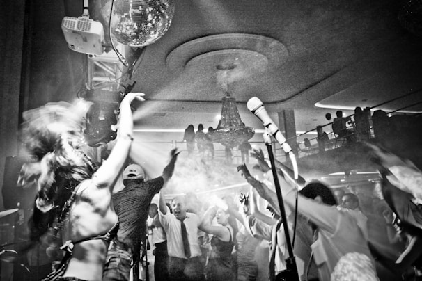 A lively party scene with a crowd of people dancing enthusiastically. The setting is a club or event venue, featuring a large chandelier hanging from the ceiling and light effects with smoke enhancing the atmosphere. At the center of the image, a person seems to be singing or speaking into a microphone on a stand. Various individuals in the foreground have their arms raised and appear to be enjoying the music.