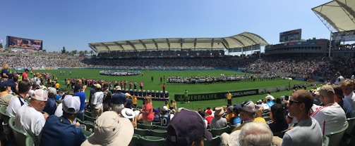 A large sports stadium filled with spectators, a well-maintained green field in the center, and a group of people forming lines as they prepare for an event. The seating areas are crowded, and the atmosphere appears lively with fans wearing various team colors and merchandise. Overhead, clear skies and bright sunlight illuminate the venue.