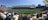 A large sports stadium filled with spectators, a well-maintained green field in the center, and a group of people forming lines as they prepare for an event. The seating areas are crowded, and the atmosphere appears lively with fans wearing various team colors and merchandise. Overhead, clear skies and bright sunlight illuminate the venue.