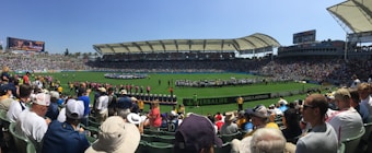 A large sports stadium filled with spectators, a well-maintained green field in the center, and a group of people forming lines as they prepare for an event. The seating areas are crowded, and the atmosphere appears lively with fans wearing various team colors and merchandise. Overhead, clear skies and bright sunlight illuminate the venue.
