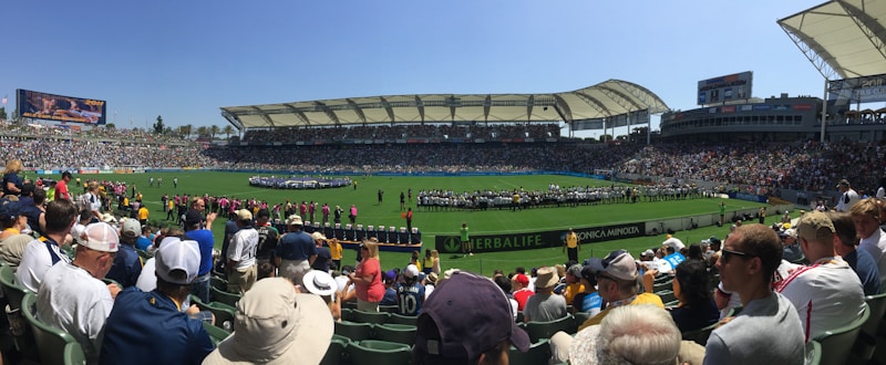 A large sports stadium filled with spectators, a well-maintained green field in the center, and a group of people forming lines as they prepare for an event. The seating areas are crowded, and the atmosphere appears lively with fans wearing various team colors and merchandise. Overhead, clear skies and bright sunlight illuminate the venue.