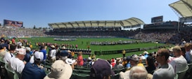 A large sports stadium filled with spectators, a well-maintained green field in the center, and a group of people forming lines as they prepare for an event. The seating areas are crowded, and the atmosphere appears lively with fans wearing various team colors and merchandise. Overhead, clear skies and bright sunlight illuminate the venue.