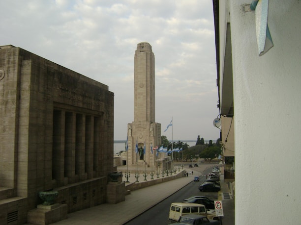 A tall stone monument is surrounded by flags, with a significant building featuring large pillars on the left side. The scene appears to be from an urban area with parked cars, people, and trees in the background. The sky is overcast, creating a somewhat muted atmosphere.