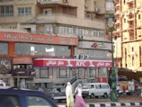 An urban street scene features a busy area with people walking past a building. This building has multiple signs in both Arabic and English, including a KFC and Little Caesars Pizza. Several vehicles, including a van and cars, are parked or moving on the street. The architecture is a mix of commercial and residential with visible air conditioning units. A lamppost is prominently featured in the foreground.