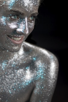A smiling person is covered in shimmering silver glitter, with some areas reflecting light in blue hues. The focus is on the face and upper body, emphasizing the sparkling texture of the glitter against a dark background.