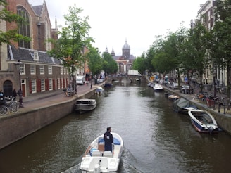A canal flanked by historic buildings and trees, with a small motorboat navigating the waterway. Bicycles are parked along the waterfront, and several people are walking or cycling on the adjacent paths. A prominent church with a large dome is visible in the background, while smaller boats are moored along the banks.