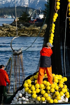 Two individuals are on a fishing boat handling large fishing nets. The man in the foreground is wearing bright orange pants and a red jacket, standing amongst a pile of yellow and white buoys. The background features more fishing boats and a rocky shoreline with snow-capped mountains.