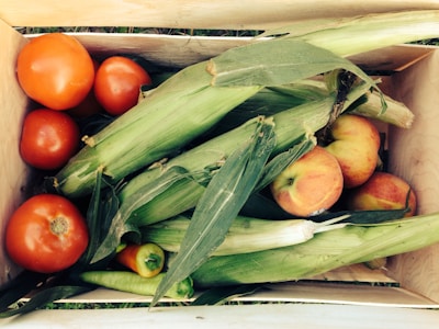 A wooden crate filled with fresh produce, including several ripe red tomatoes, ears of corn with husks, and a few peaches. Nestled among the vegetables is a small pepper. The arrangement showcases a variety of textures and natural colors.
