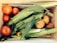 A wooden crate filled with fresh produce, including several ripe red tomatoes, ears of corn with husks, and a few peaches. Nestled among the vegetables is a small pepper. The arrangement showcases a variety of textures and natural colors.