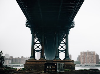 An urban scene captures a bridge from underneath, showcasing its massive steel framework. In the foreground, a dumpster with a company name and phone number is visible. The background consists of high-rise buildings under a cloudy sky, suggesting an overcast day.