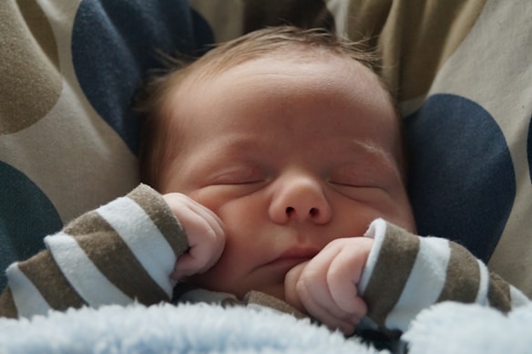 A newborn baby peacefully sleeping with closed eyes, wearing a brown and white striped outfit. The infant's hands are gently tucked under the chin. The background includes a soft cushion with blue and beige polka dots.