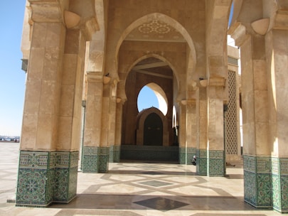 Arched stone structures with ornate green tile patterns on the lower sections create a symmetrical and grand architectural design. The flooring also features intricate geometric patterns. Natural light filters through the arches, highlighting the textures and colors.