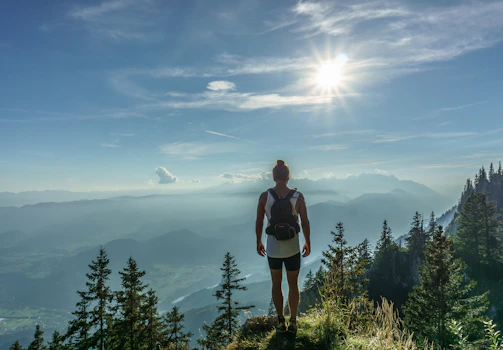 A traveler with a backpack standing on a mountain trail overlooking a sunrise landscape.
