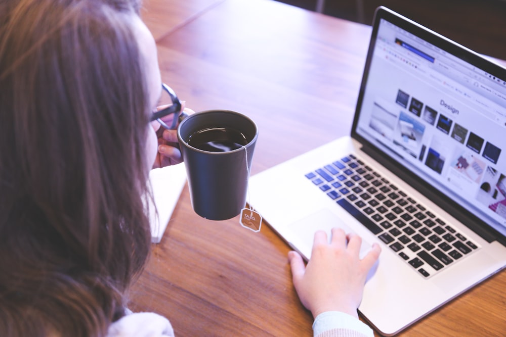 mujer sosteniendo una taza llena de té usando MacBook