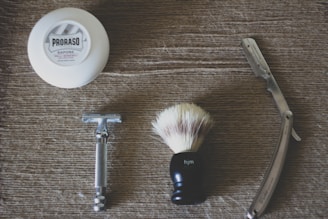 A flat lay of shaving essentials on a textured surface, including a white tub of shaving soap labeled 'Proraso', a metal safety razor, a shaving brush with a black handle, and a straight razor with a shiny blade.