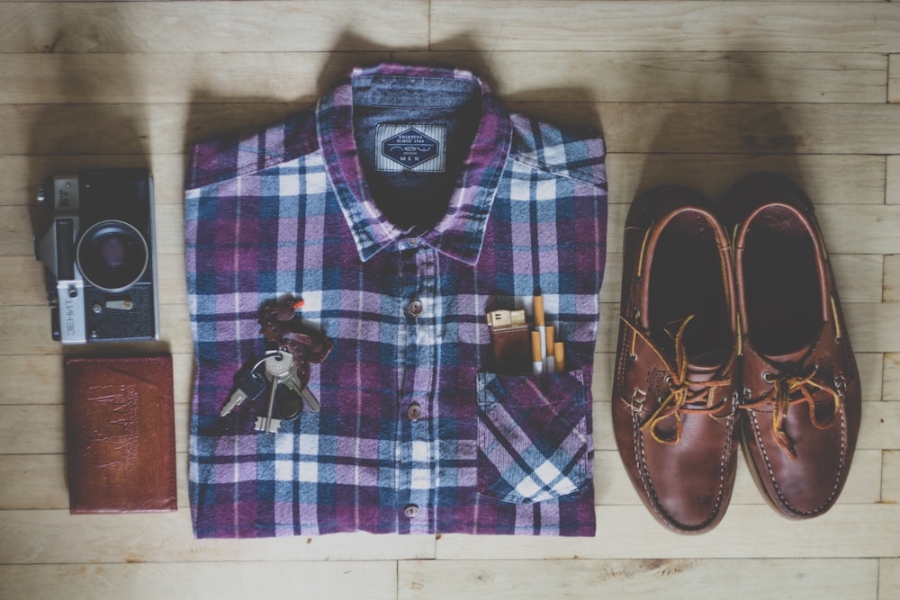 Fotografía plana de camisa abotonada, cámara, llaves, cigarrillos y un par de zapatos náuticos de cuero marrón