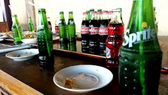 Several bottles of soda, including Sprite and Coca-Cola, are arranged on a wooden table alongside empty plates. The scene suggests a casual dining or party setting, with straws in some bottles and remnants of food on the plates.