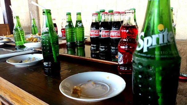 Several bottles of soda, including Sprite and Coca-Cola, are arranged on a wooden table alongside empty plates. The scene suggests a casual dining or party setting, with straws in some bottles and remnants of food on the plates.