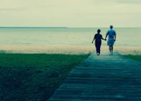 A cinematic photo of a couple walking hand in hand along a sandy path with charcoal shadows.