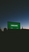 A large outdoor movie screen displays a preview notice against a darkening sky at dusk. Silhouettes of trees and landscape are visible at the bottom, and the sky transitions from a deep blue to dark gray.