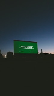A large outdoor movie screen displays a preview notice against a darkening sky at dusk. Silhouettes of trees and landscape are visible at the bottom, and the sky transitions from a deep blue to dark gray.