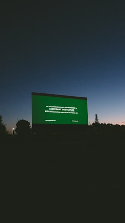 A large outdoor movie screen displays a preview notice against a darkening sky at dusk. Silhouettes of trees and landscape are visible at the bottom, and the sky transitions from a deep blue to dark gray.