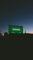 A large outdoor movie screen displays a preview notice against a darkening sky at dusk. Silhouettes of trees and landscape are visible at the bottom, and the sky transitions from a deep blue to dark gray.