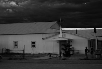 A rural church with a metal roof sits beneath a dramatic, cloudy sky. The building is simple with a white exterior and a cross on the roof. In front of the church, two children are engaged in activity, possibly playing on the ground. A basketball hoop is visible on the right, suggesting a community setting.