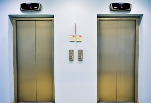 Two closed elevator doors are positioned side by side, set against a light-colored wall. Above each elevator is a digital display showing the letter 'L'. Between the elevators are control panels with buttons, and there are two small notes attached to the wall, each with visible text.