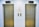 Two closed elevator doors are positioned side by side, set against a light-colored wall. Above each elevator is a digital display showing the letter 'L'. Between the elevators are control panels with buttons, and there are two small notes attached to the wall, each with visible text.