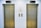 Two closed elevator doors are positioned side by side, set against a light-colored wall. Above each elevator is a digital display showing the letter 'L'. Between the elevators are control panels with buttons, and there are two small notes attached to the wall, each with visible text.
