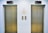 Two closed elevator doors are positioned side by side, set against a light-colored wall. Above each elevator is a digital display showing the letter 'L'. Between the elevators are control panels with buttons, and there are two small notes attached to the wall, each with visible text.