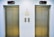 Two closed elevator doors are positioned side by side, set against a light-colored wall. Above each elevator is a digital display showing the letter 'L'. Between the elevators are control panels with buttons, and there are two small notes attached to the wall, each with visible text.