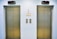 Two closed elevator doors are positioned side by side, set against a light-colored wall. Above each elevator is a digital display showing the letter 'L'. Between the elevators are control panels with buttons, and there are two small notes attached to the wall, each with visible text.